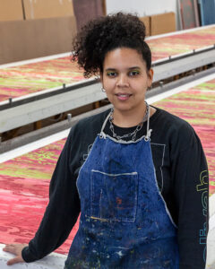 A portrait of the artist Joiri Manaya, an Afro-Latino woman wearing a blue apron over a long-sleeve black shirt. She is standing in front of printing tables in a studio, with colorful stretches of yardage laid out atop the tables.