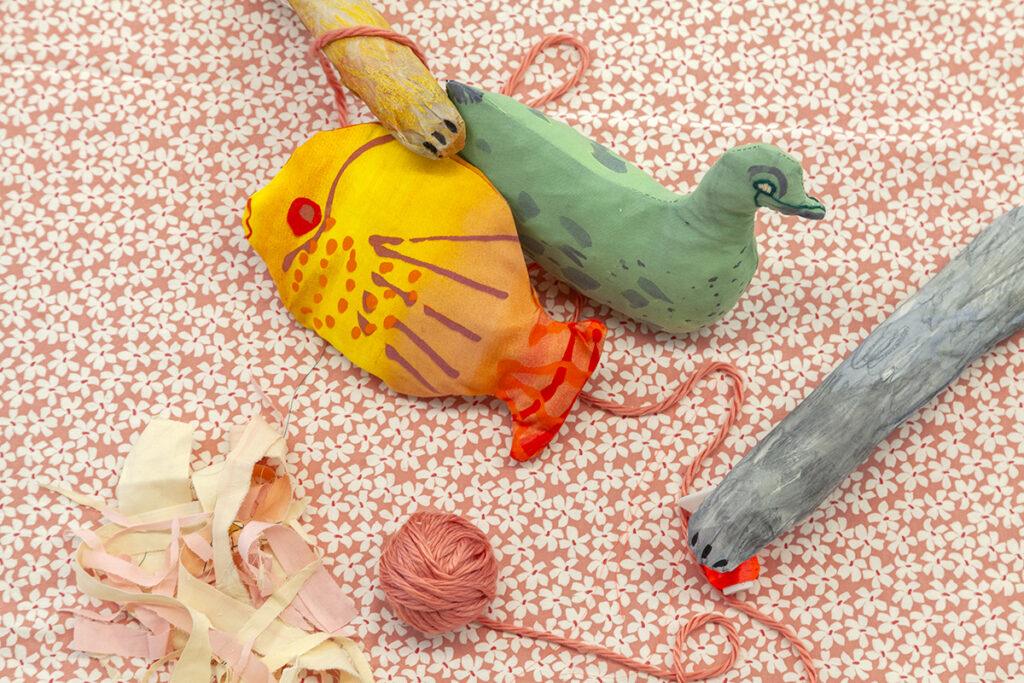 A photo of stuffed animals, including a yellow fish, a green duck, and two cat paws entering the frame to paw at the fish and a ball of twine.