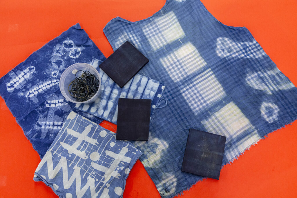 An assortment of fabrics (napkins, bandanas, shirt) that have been dyed in indigo using a shibori technique showing off different dye patterns.