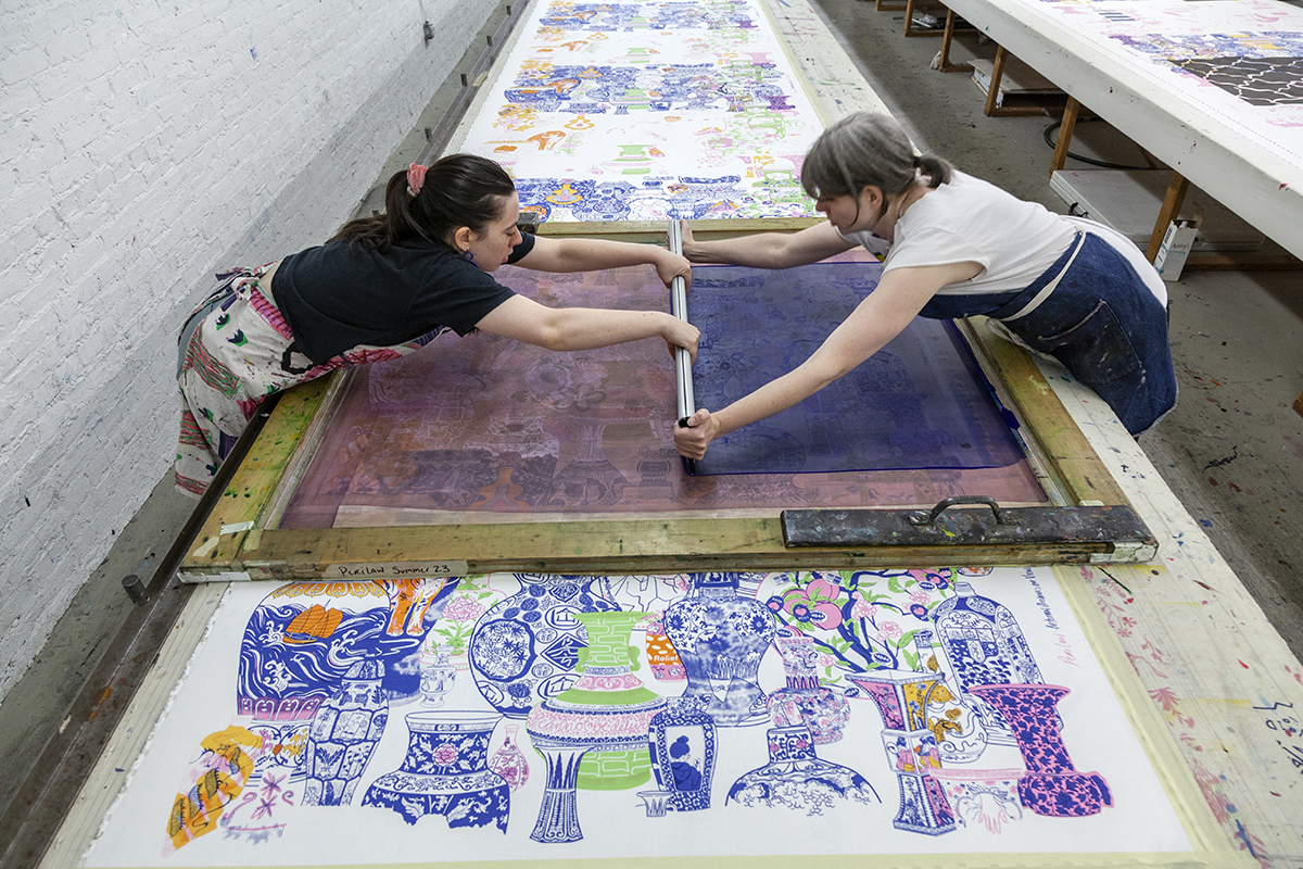 Two women work together to screen print on a very long table. Positioned one on each side of the table, they stretch their arms across it to grip a squeegee and move ink across a silkscreen. They are printing a pattern consisting of East Asian-style vases, with decorative patterns and pictures on them.