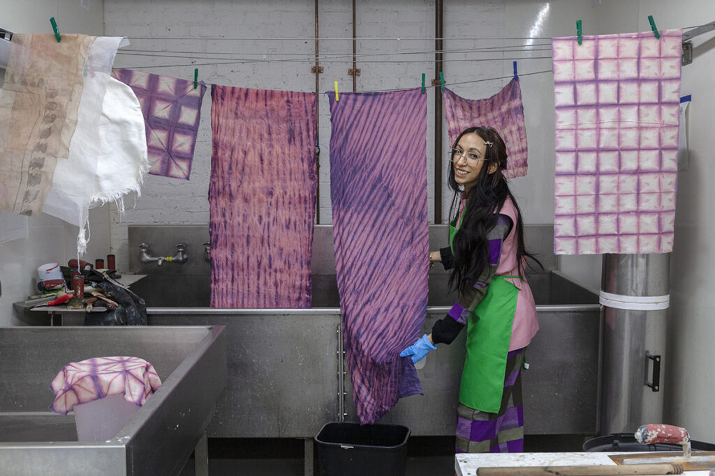 The artist Lisa Alvarado, a Chicanx woman with long straight dark hair, smiles next to an arrangement of dyed textiles on a clothesline over a large industrial sink.