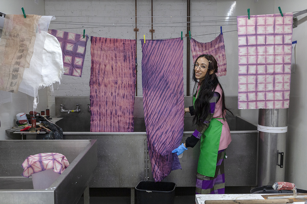 The artist Lisa Alvarado, a Chicanx woman with long straight dark hair, smiles next to an arrangement of dyed textiles on a clothesline over a large industrial sink.