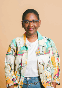 A portrait of Keyonna Butler, a young Black woman wearing glasses and a colorful jacket over a white t-shirt. 