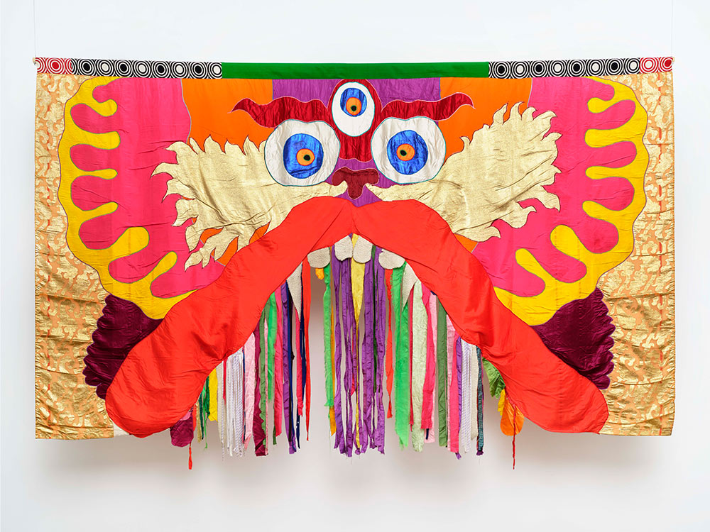 A photo of a fabric-based artwork of the face of a three-eyed dragon staring directly at the viewer. The face is cartoonish and mostly symmetrical, with different colors of fabric emanating outwardly from its nose. It has a large red upper lip that bows upward, with colorful tassels hanging under its teeth.