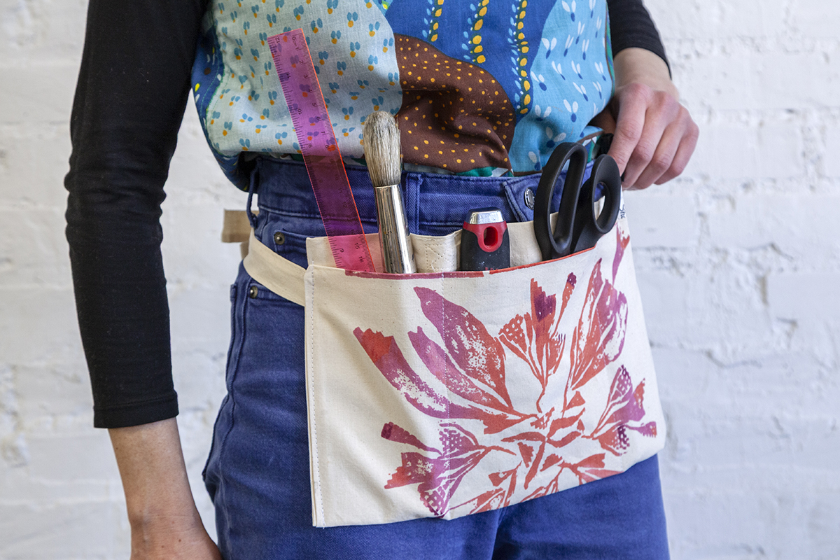 A photo of a canvas tool belt worn at someone's waist. A ruler, paintbrush, utility knife, and a pair of scissors are tucked in the pockets.