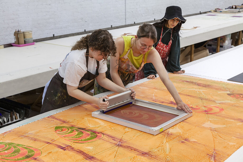 Three studio artists screenprint a detail over a large printed cloth while Lisa Alvarado, an Artist-in-Residence observes.