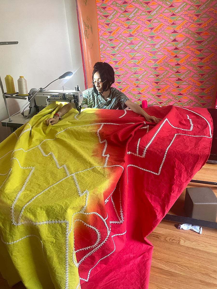 An artist uses a sewing machine on a large tapestry in her studio. The fabric is split dyed yellow and red and overflows beyond her table.