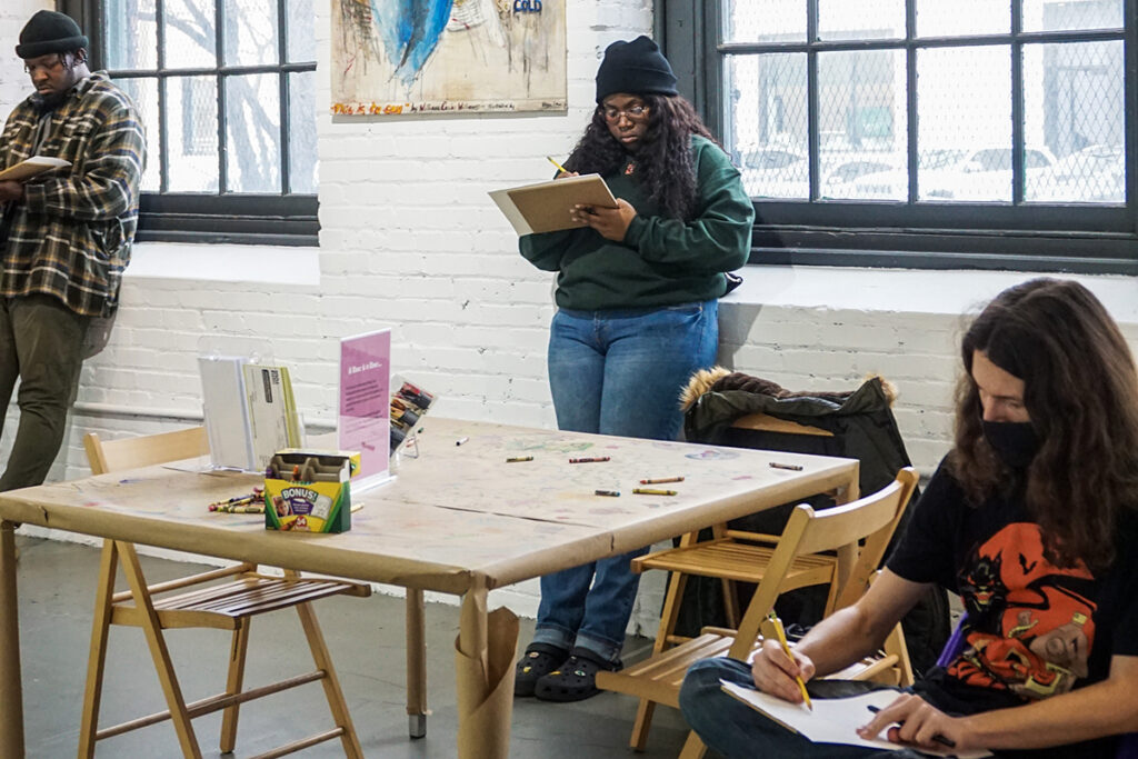 A group of young adults spread out standing and sitting around a table and facing the same direction while drawing in their sketchbooks.