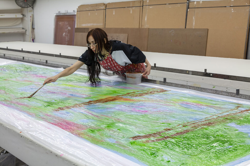 The artist Lisa Alvarado, a Chicanx woman with long straight dark hair and glasses, leans over a large print table with a brush. The table's surface features a wet, mostly green sheet of fabric covered in plastic.