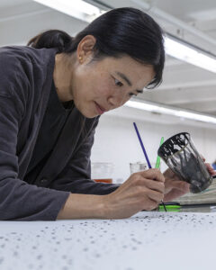 A photo of the artist Julia Chiang, an Asian-American woman, painting with a fine-tipped brush on a surface.