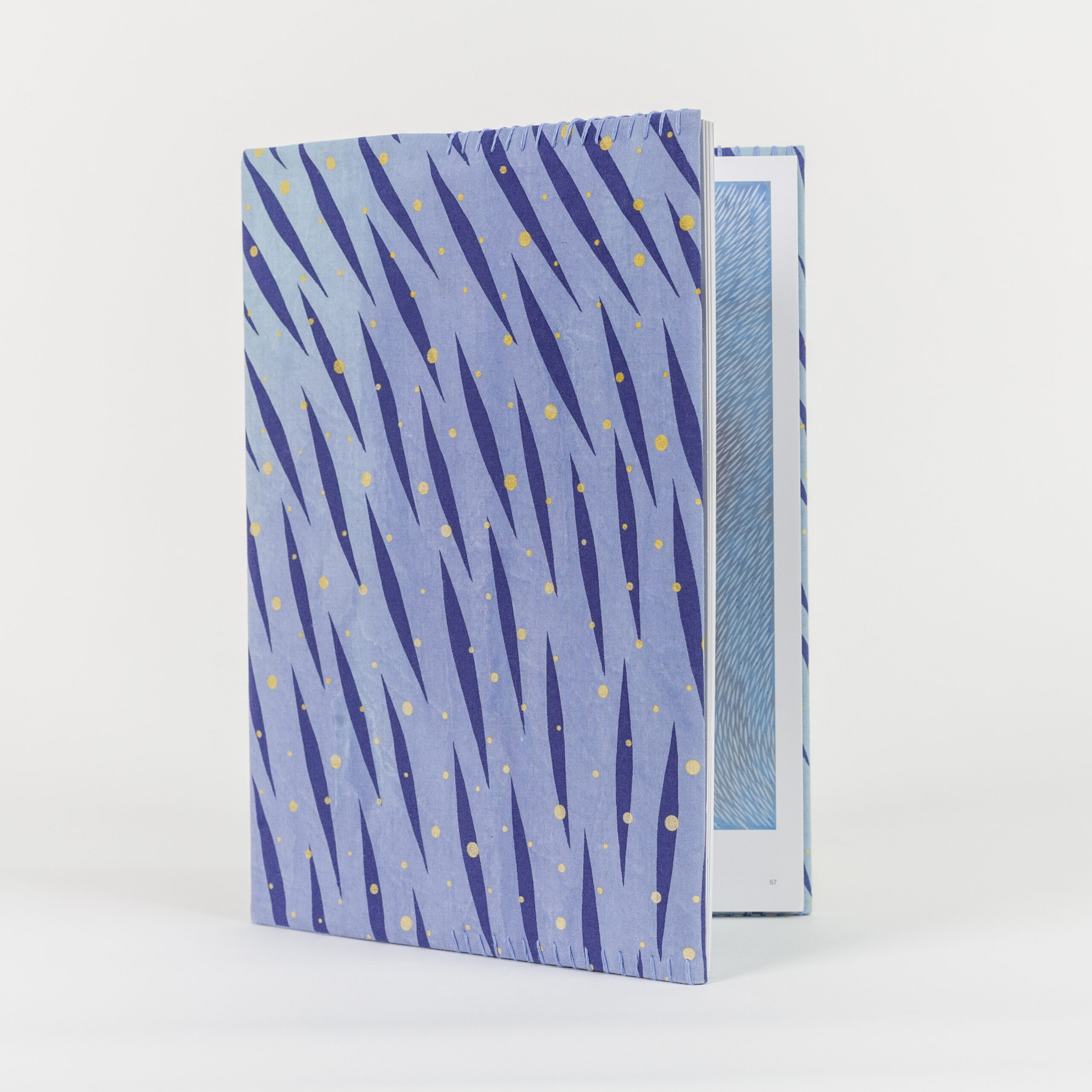 A photo of a hardback book with a hand-stitched book cover featuring a vibrant, pattern of diagonal dark blue brush-like streaks and scattered yellow dots on a light blue background. The book is slightly open, revealing a blue patterned illustration inside.