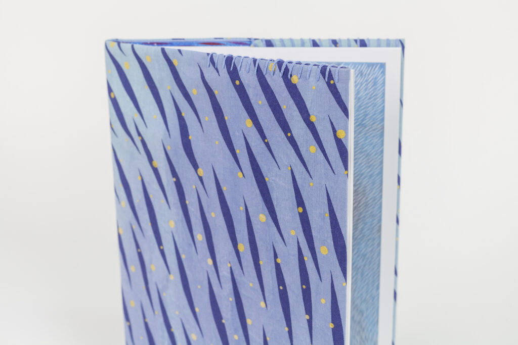 A photo of a hardback book with a hand-stitched book cover featuring a vibrant, pattern of diagonal dark blue brush-like streaks and scattered yellow dots on a light blue background. The book is slightly open, revealing a blue patterned illustration inside.