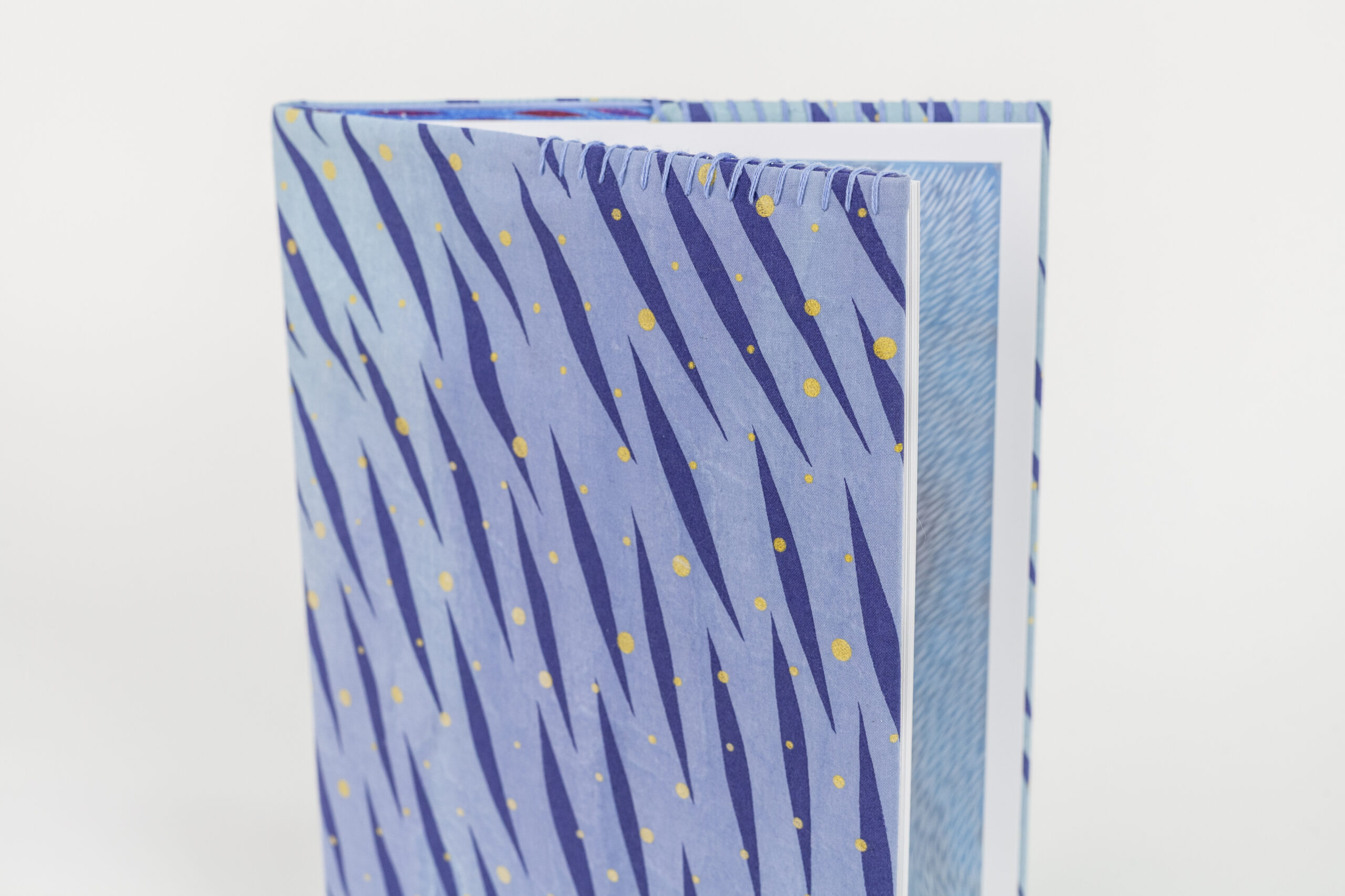 A photo of a hardback book with a hand-stitched book cover featuring a vibrant, pattern of diagonal dark blue brush-like streaks and scattered yellow dots on a light blue background. The book is slightly open, revealing a blue patterned illustration inside.