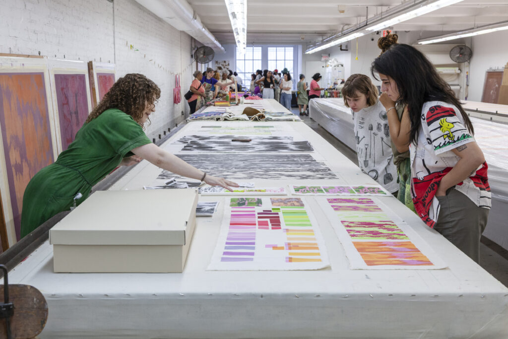 A photograph of three people leaning over a table with screenprinting process materials displayed and organized on top. One person on the left has their arm stretched over the table pointing at the materials and the two people on the right are listening and observing.