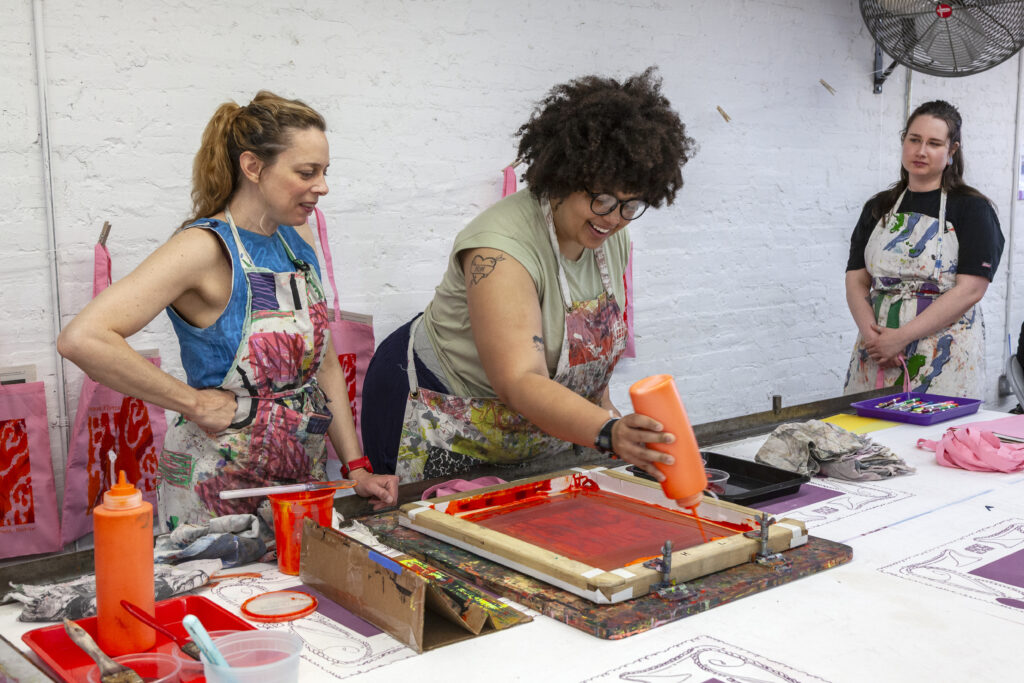 Three people gathered around a table watching one person screenprint on the table top.