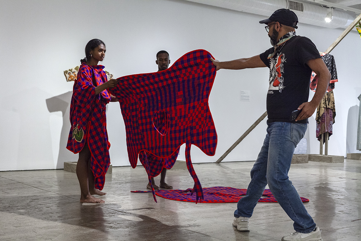 The artist Brendan Fernandes pulls on a red and blue plaid costume pieces held by a dancer who is wearing the same garment.