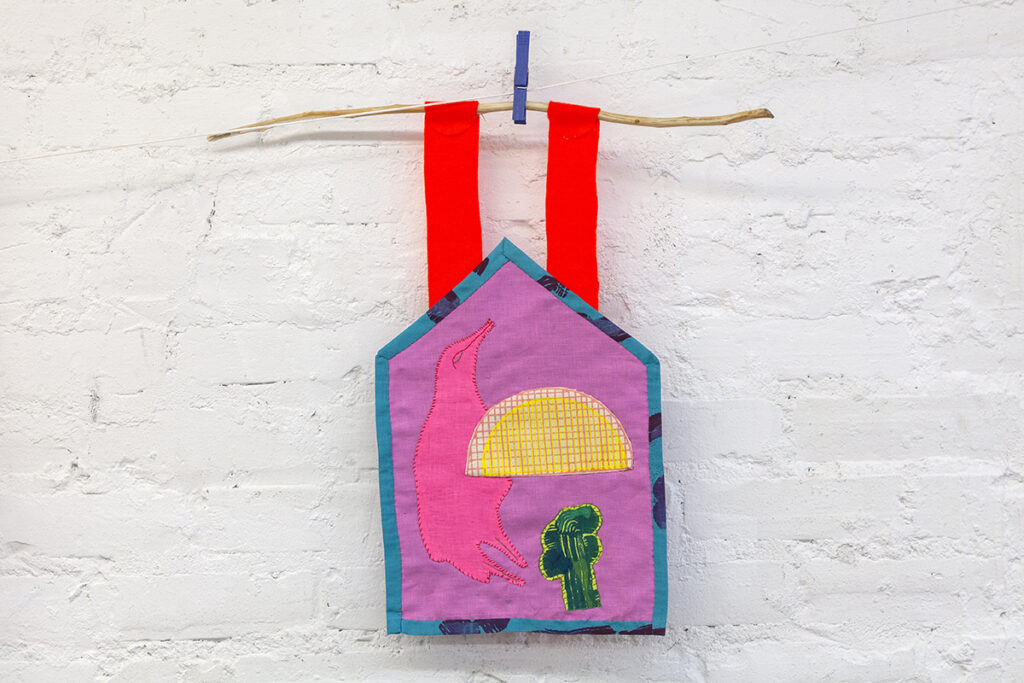 A hand sewn small fabric tapestry featuring an appliqué motif of a bird and a sunset. The tapestry is hanging from a small branch against a painted brick wall.