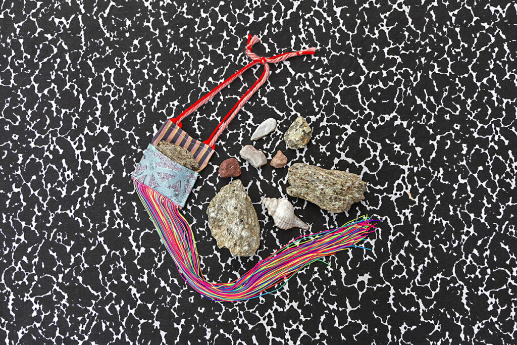 A colorful hand sewn pocket bag for a small rock lays on a composition pattern background next to an assortment of different types and sizes of rocks and pebbles.