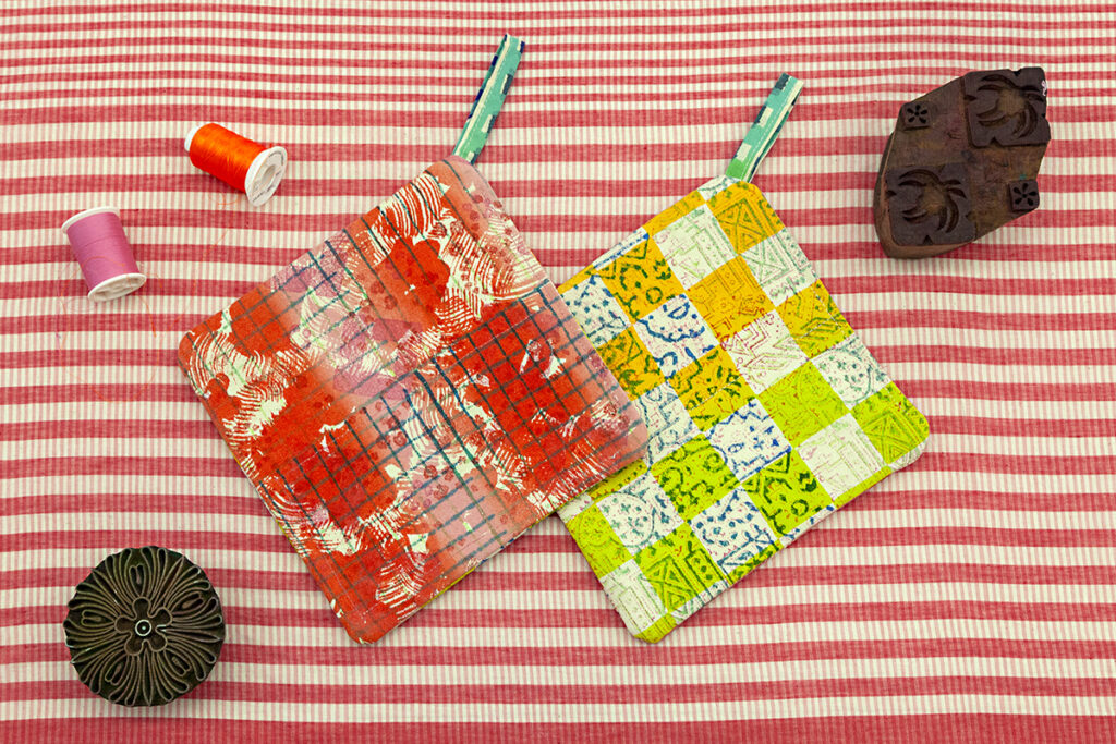 A pair of colorful patterned potholders laying on red and white striped fabric. Hand-carved woodblocks and spools of thread are surrounding the potholders.