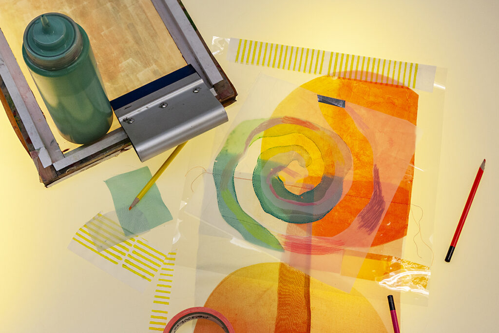 Samples of screenprints on transparent mylar and scraps of transparent fabric displayed on a light table emitting a soft warm light.