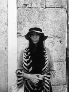 A photo of the artist Lisa Alvarado, a woman with long dark hair and glasses. She leans against a wall outdoors while wearing a sun hat and a long scarf.