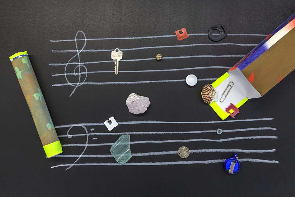 Different pieces of forgotten found objects like keys, bread ties, rocks, paper clips and more spread out as if they are falling out of a screenprinted paper box onto a drawn illustration of a blank sheet of music.