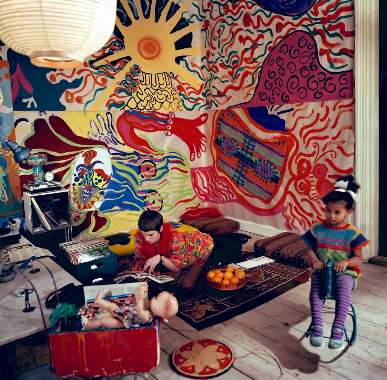 An older photograph from the 1960s of a woman and her young daughter in a living room. The woman is lounging on the floor reading a book while the little girl, seated on a rocking horse, is throwing an object. There is a large, colorful hand-painted mural on the wall, a record player, record crate, and toys nearby.