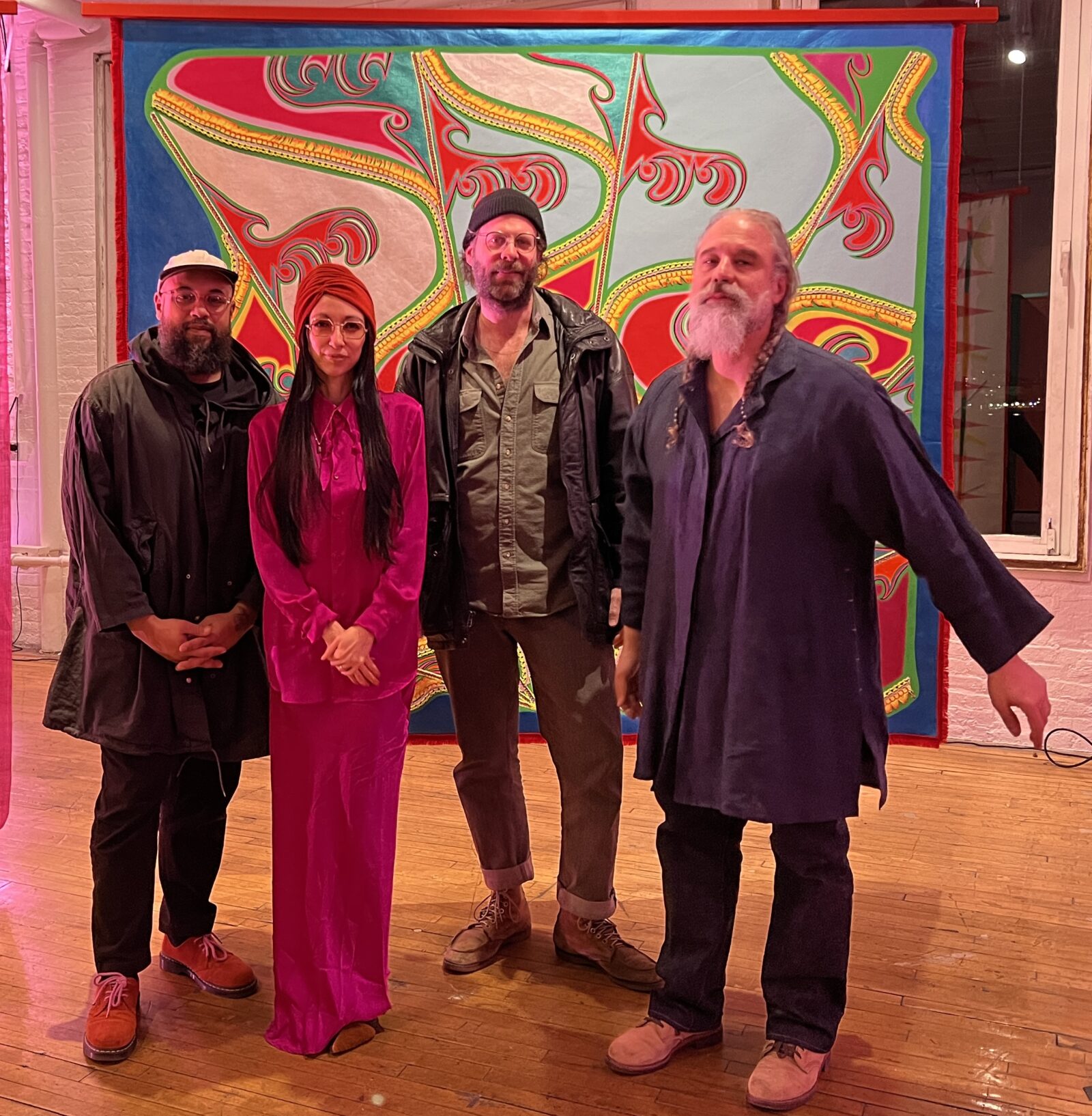A group of four people standing together in a magenta-lit room. They are standing in front of an abstract and colorful handmade tapestry.