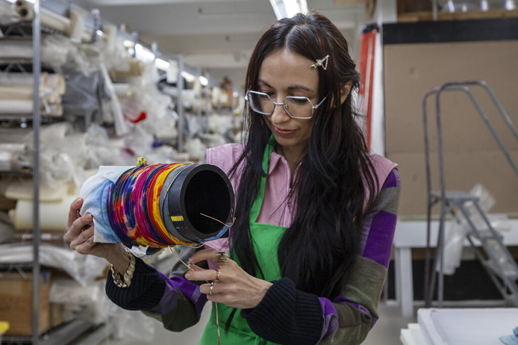 Artist Lisa Alvarado is standing in a studio with shelves of art and fabric materials in the background. She is holding a spool of colorful fabric wrapped around a cone.