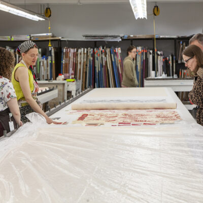 A group of adults standing around a large fabric printing table in a studio looking at decorative, patterned fabric that is laid out across the table.