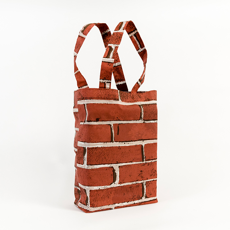 A photo of a tote bag pictured at a slight angle against a white background. The bag features a red brick pattern wrapped around all sides. Two thin handles stand in an upright loop from each side.