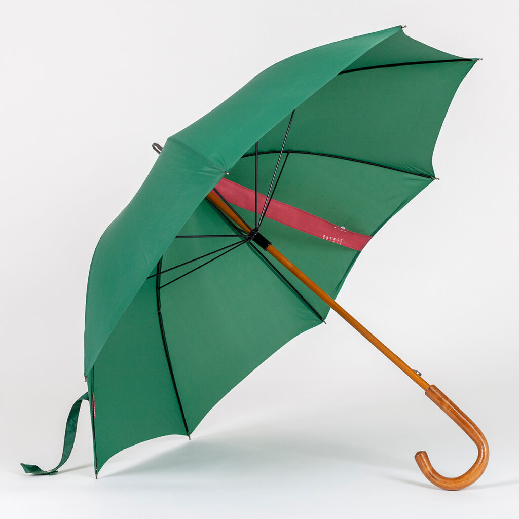 A green umbrella with a tan wood handle, supported by a black metal armature. A red strip of fabric stretches from its center toward its outer band, acting as a sundial.