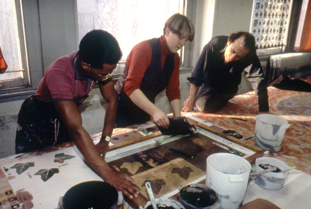Three people lean over a table, all focused on art being printed. The woman at the center is lifting a squeeguee from the table as the man on the left holds down a silkscreen.