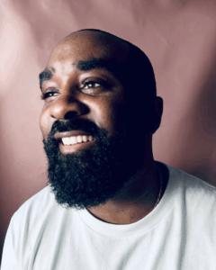 A portrait of the artist and creative director Shaeed Rucker. A Black man with a shaved head and thick goatee, Rucker wears a white t-shirt and smiles, turned at three-quarters.