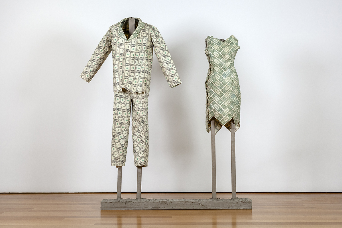 Two garments, a man's suit and a woman's dress, are installed on armatures in a gallery. The outfits are constructed entirely of US one-dollar bills.