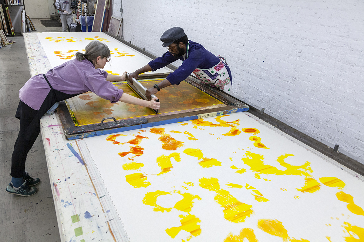A photo of two artists stretched across each side of a long table to work together in screenprinting one layer of a repeat pattern.
