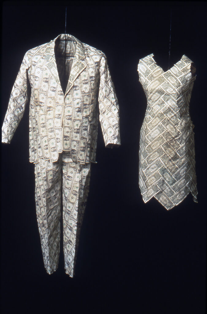 Two garments, a man's suit and a woman's dress, float in a black void. The outfits are constructed entirely of US one-dollar bills.