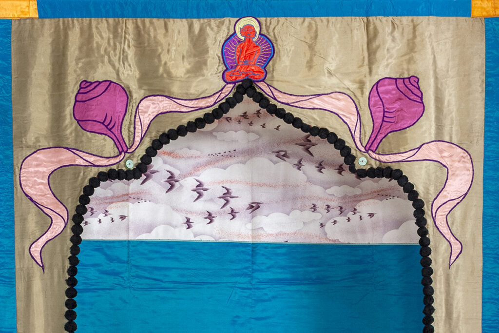 A fabric tapestry with a decorative archway shape. There is a person sitting cross-legged in the center atop the archway. Through the arch, there is a fabric with a pattern of birds flying in a cloudy sky above a plain blue fabric.