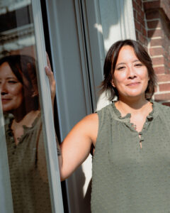 A photograph of poet Jericho Jennifer Nelson standing outside next to a window where you can see their reflection in the window.
