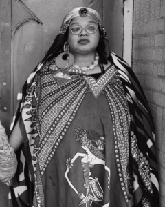 A portrait of Angel Bat-Dawid, a woman of color who is wearing decorative and powerful attire. She is standing in front of a doorway.