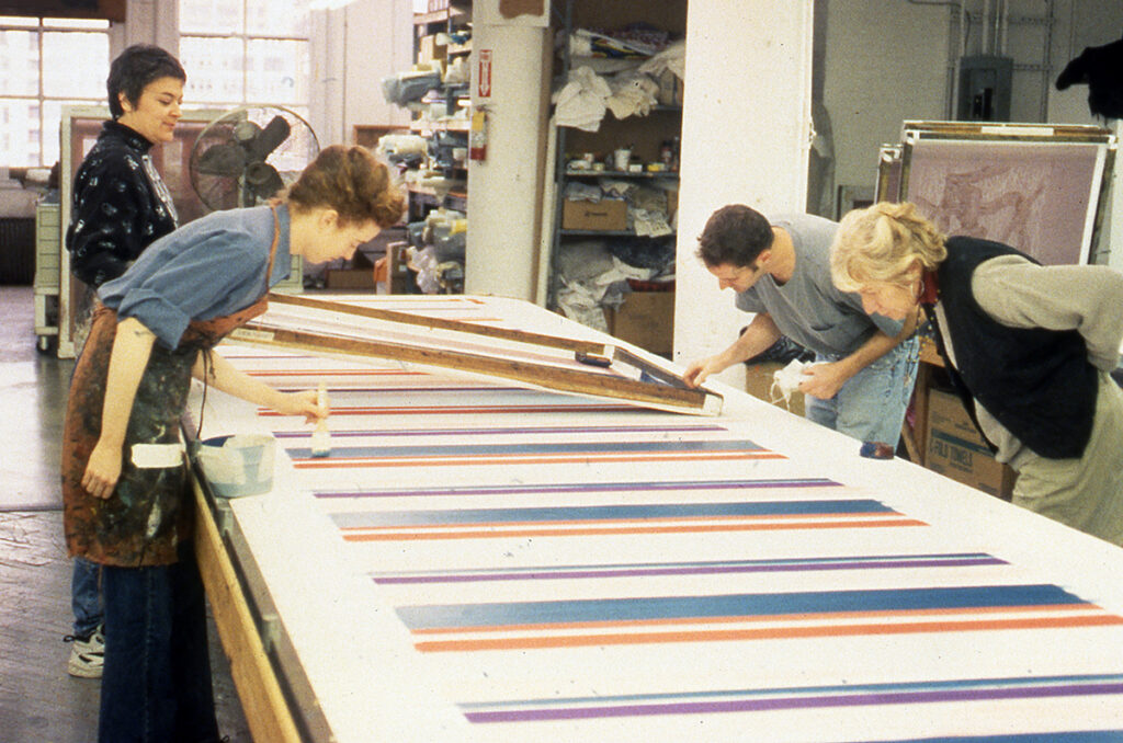Artists working in a studio examine screenprints of colorful stripes on a long printmaking table.