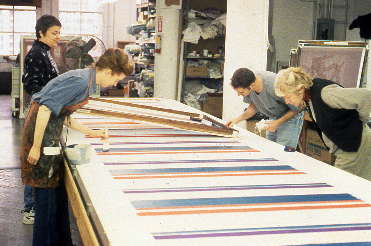 Artists working in a studio examine screenprints of colorful stripes on a long printmaking table.