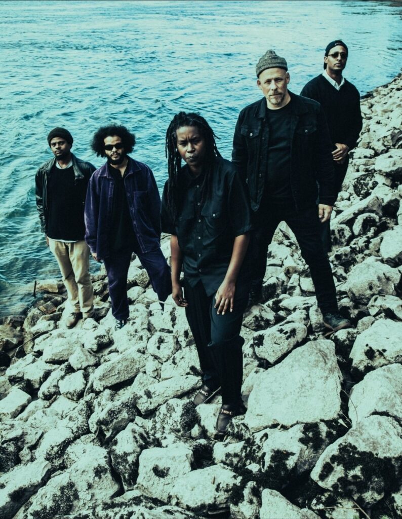 A photo of the band Irreversible Entanglements, a quintet of musicians seen standing on a rocky shore.