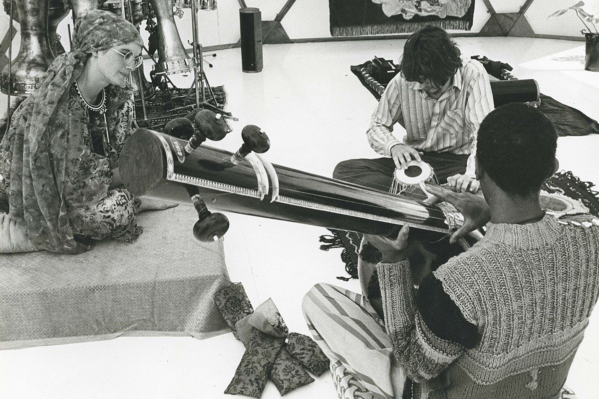 A black and white photograph of musicians sitting on the floor with instruments and music-making equipment around them.