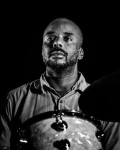 A portrait of the jazz percussionist Chad Taylor. Taylor, a Black man with a beard, looks out to an audience from his drum kit.