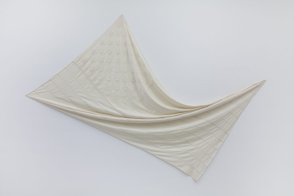 A photo of an artwork in the form of a white desaturated American flag pinned to a wall in a way that twists it from corner to corner.