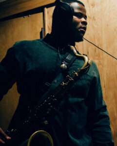 A photograph of the musician Aaron Shaw seen in three-quarters. He is a Black man wearing headphones and a long-sleeve button-up shirt, holding his saxophone.