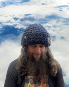 A photograph of the musician Carlos Niño, smiling slightly while glancing downward. He is a white man with long hair and a long beard wearing a t-shirt and a large knitted beanie.