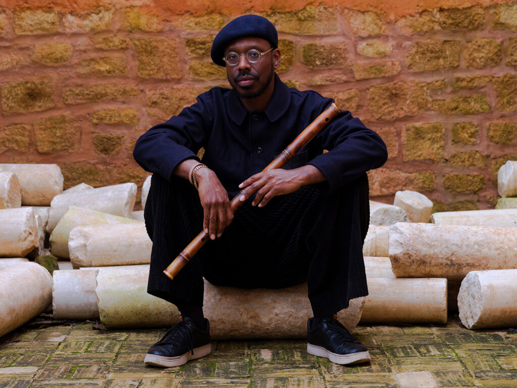 A photo of the musician Shabaka Hutchings, a Black man with a flat cap and a flute. He is seated outside on broken pillars of concrete.