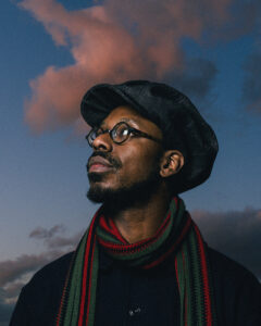 A profile portrait of the musical artist Shabaka, a Black man in a flat cap, glasses, and a scarf. He is pictured outside with a warm-toned cloud behind him. 
